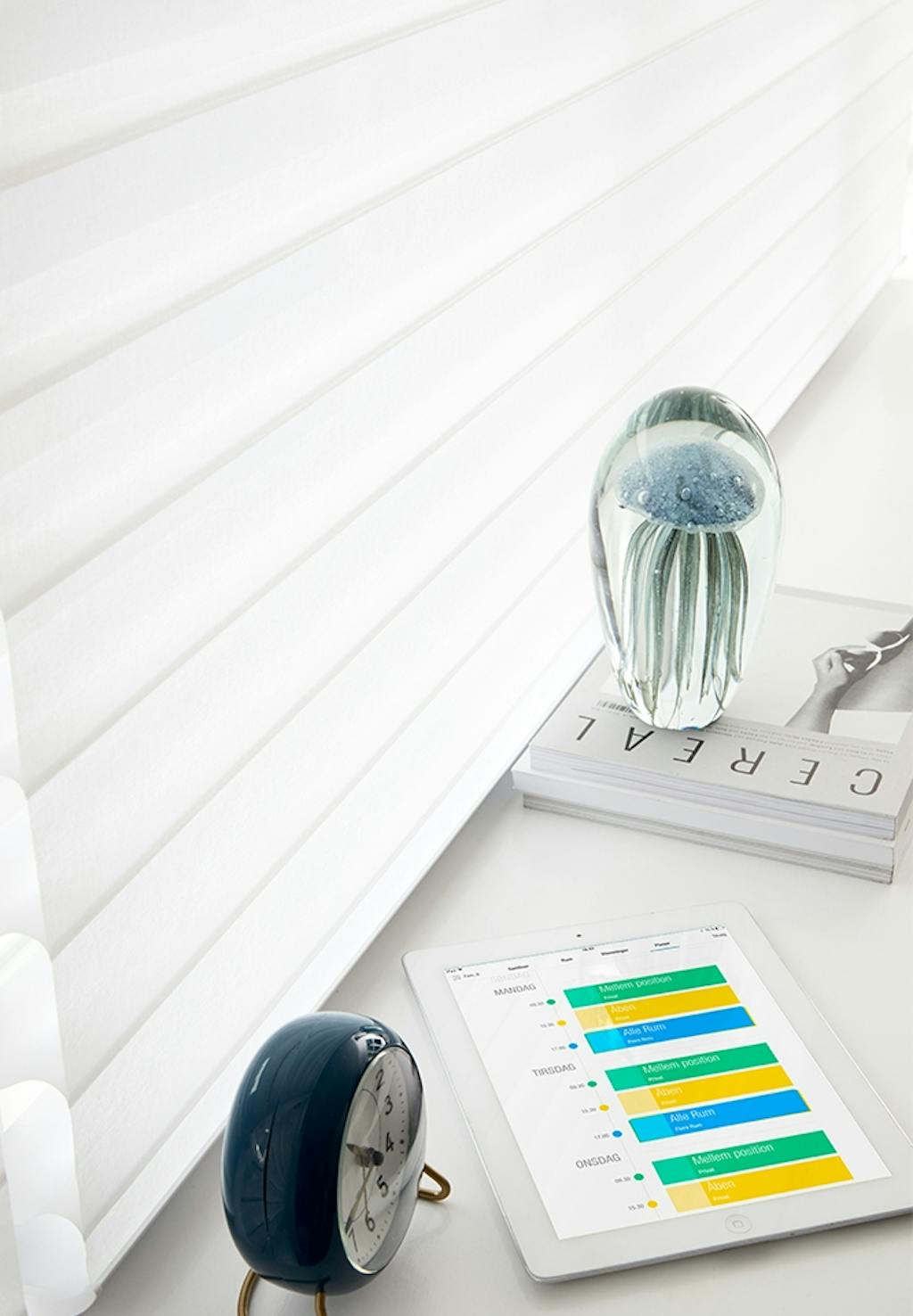 Vækkeur, iPad og bog i vindueskarm foran hvidt gardin.
