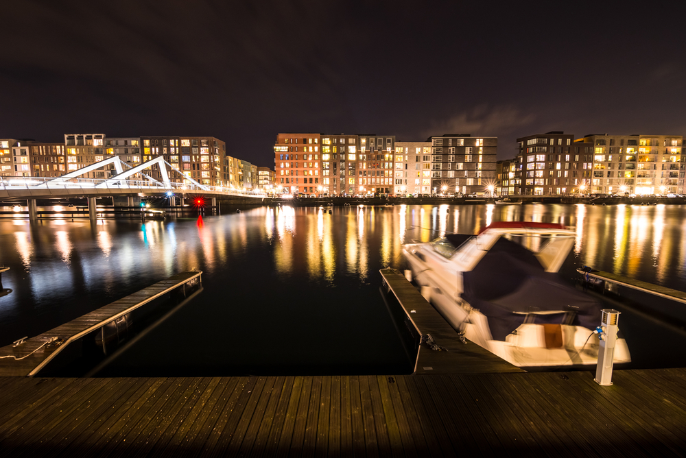 Båd ligger i dok foran lysende bylandskab hvor det er fotograferet med lang lukkertid og varm hvidbalance