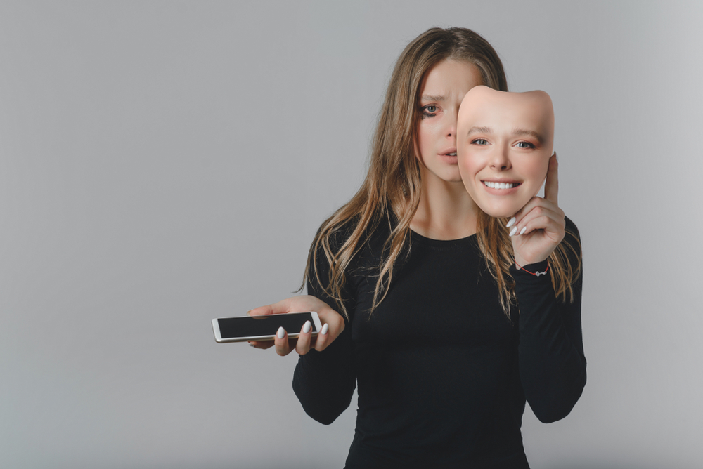 Kvinna med mobiltelefon som håller upp leende mask framför ansiktet