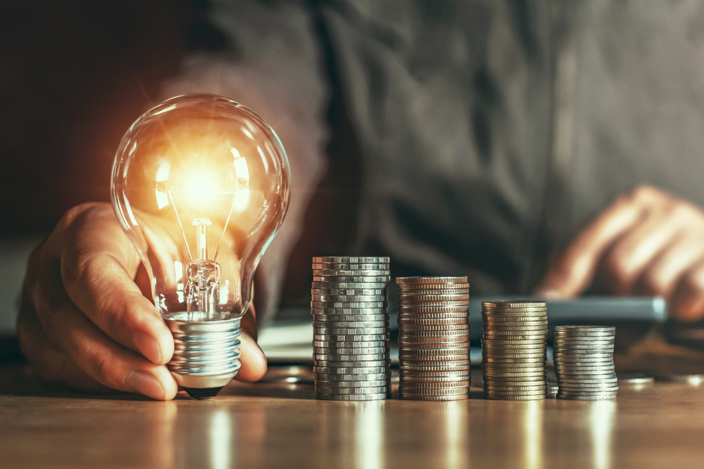 person holder lysende pære frem mod kameraet ved siden af stakkevis af sparede penge