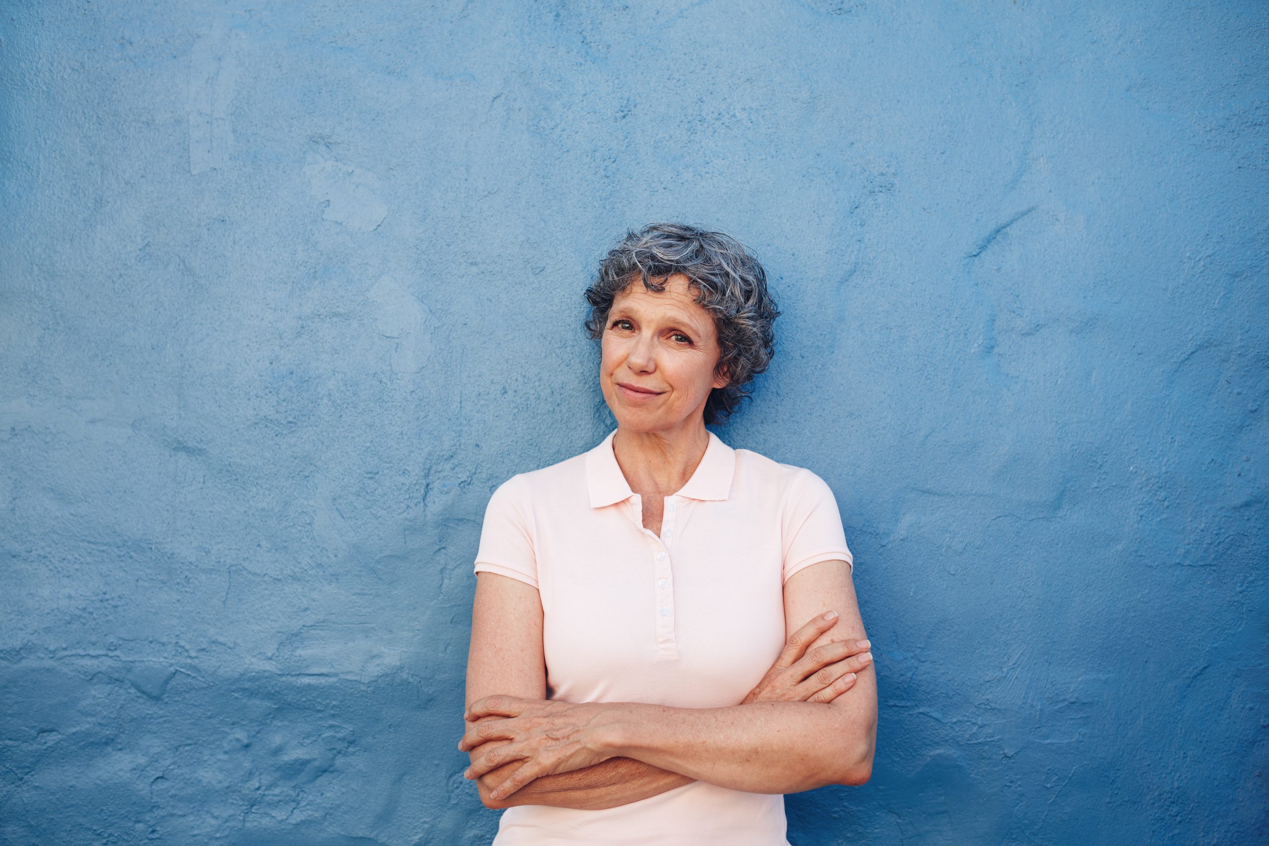 Vanhempi nainen seisoo kädet puuskassa sinistä seinää vasten