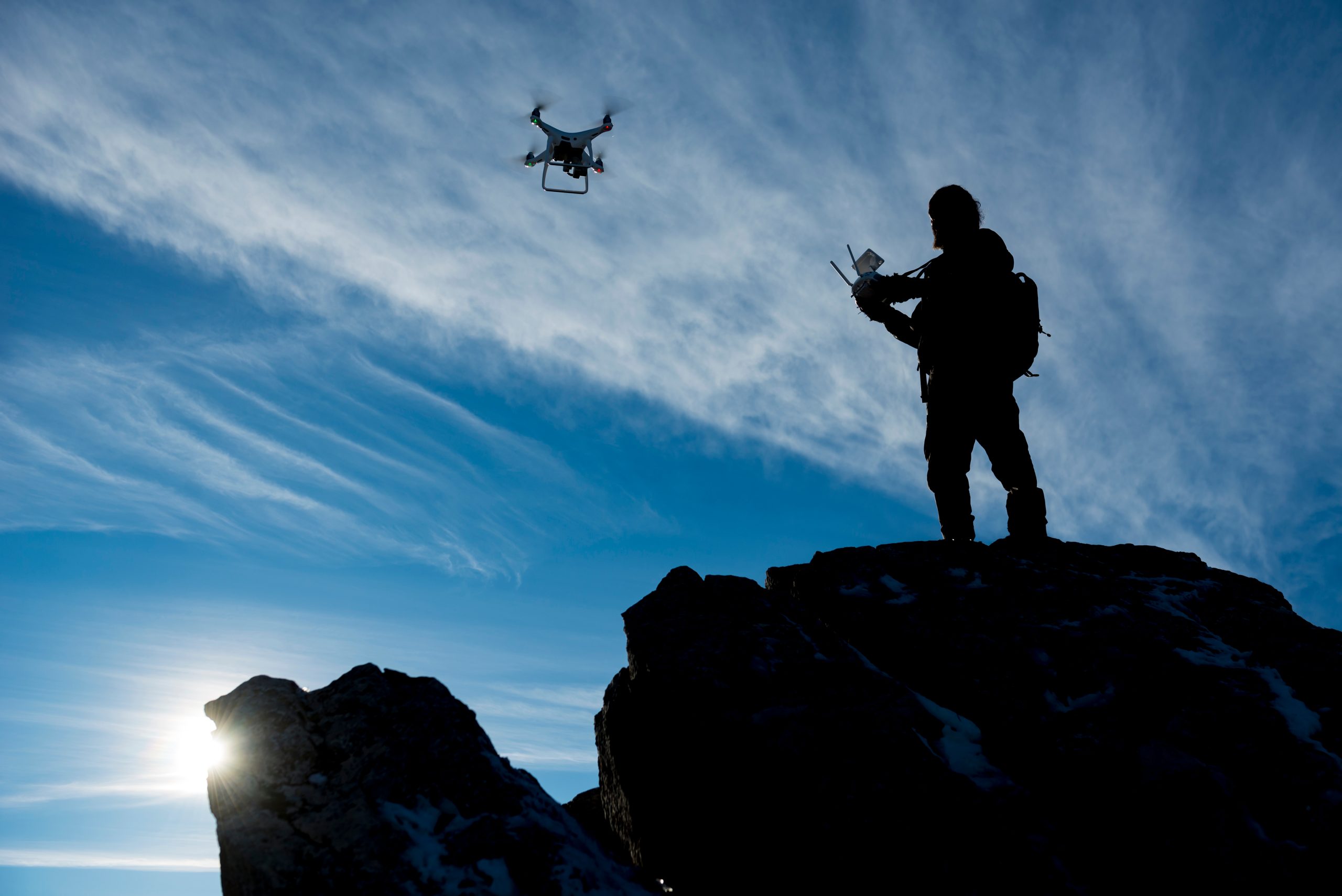 Drone og dronepilot ses som silhuetter mot blå himmel og motlys fra solen