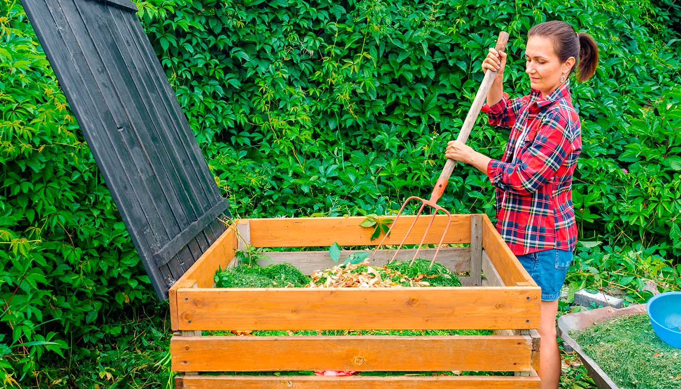 Kvinne bruker en høygaffel til å blande kompost i en kompostbeholder av tre i en hage med grønt løvverk.
