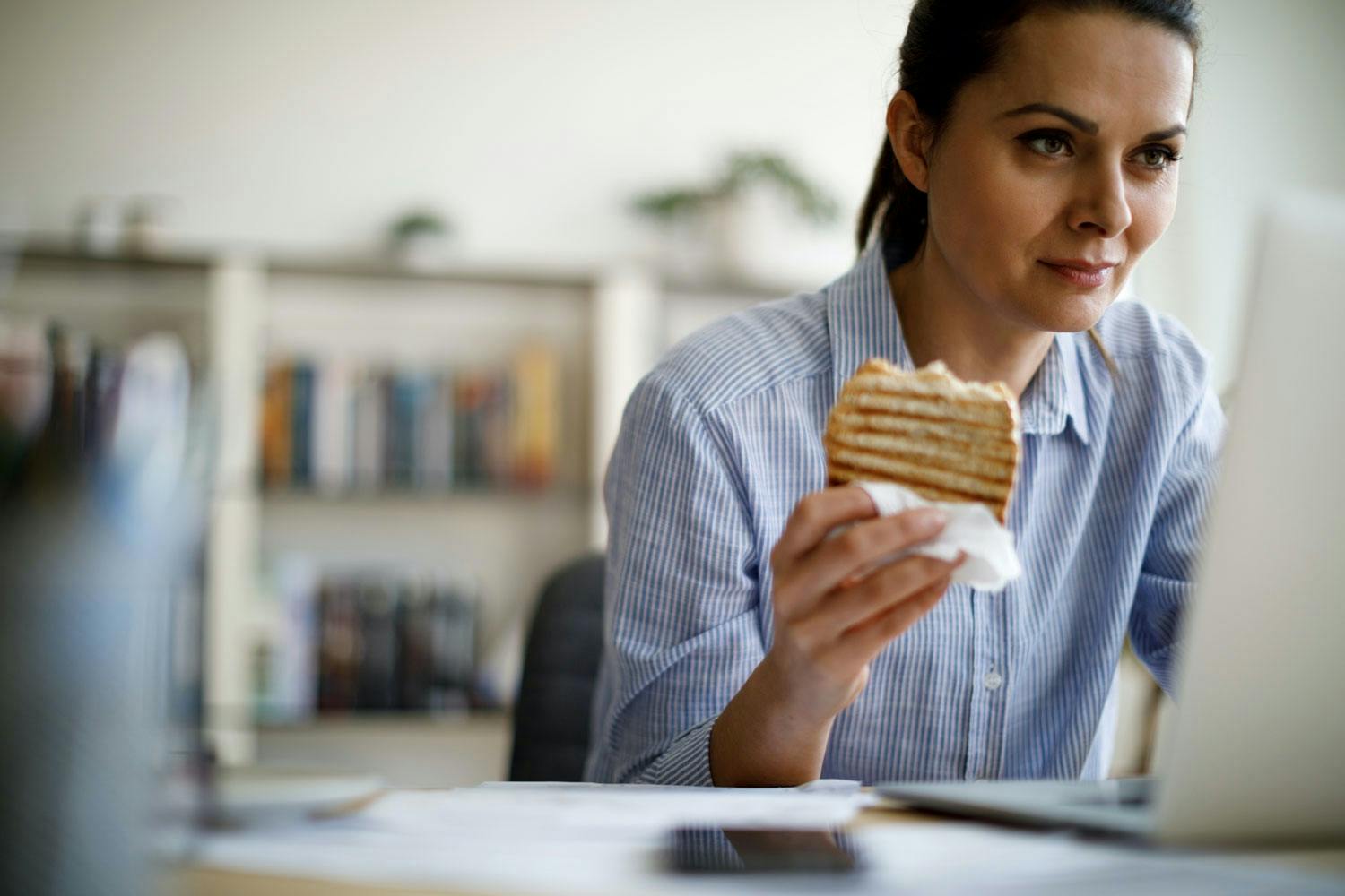 Kvinne som spiser usunt foran PC-en på jobben. 