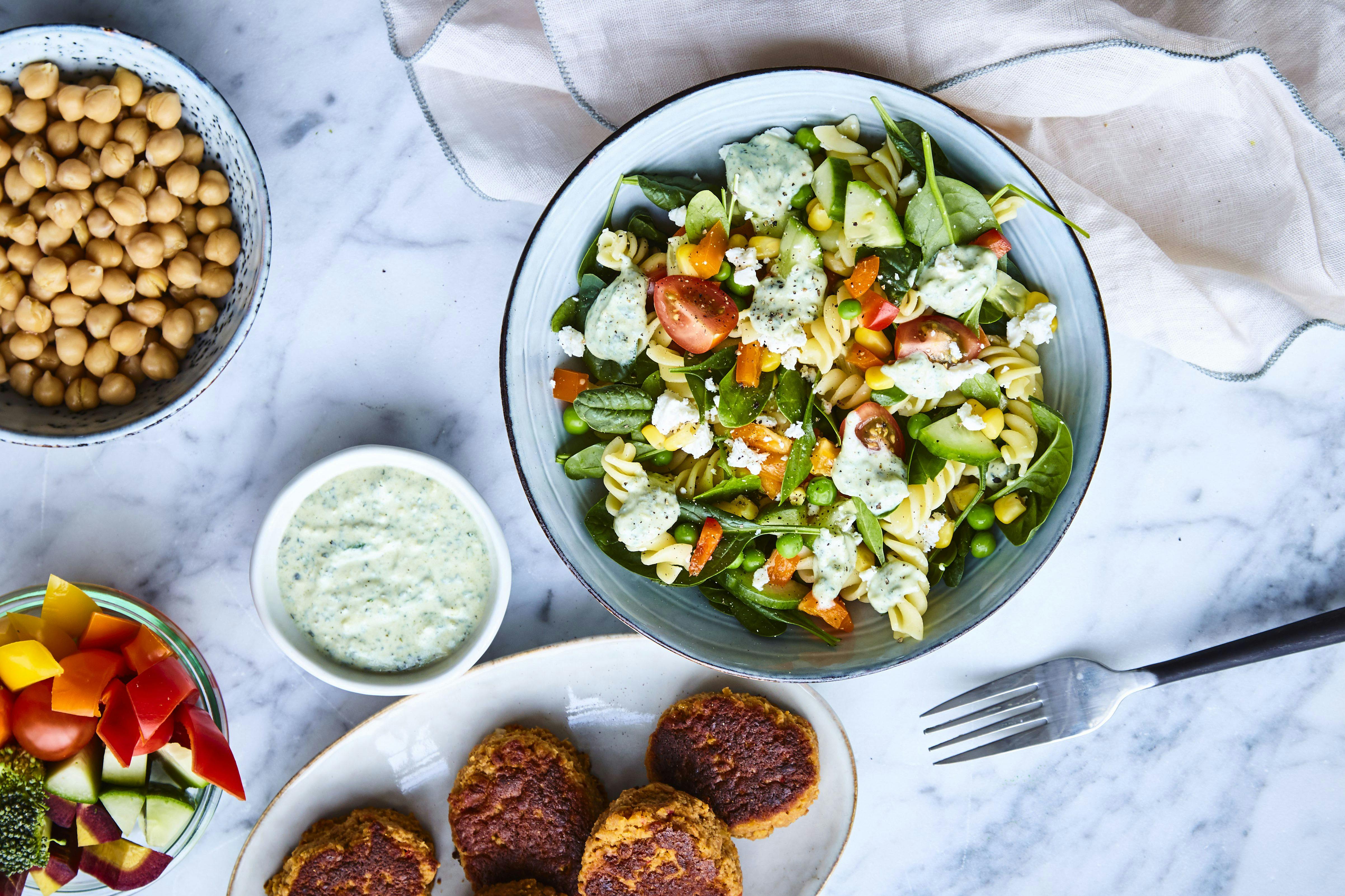 Tallerken med vegetarkaker, tallerken med pastasalat, tallerken med kikerter, tallerken med grønnsaker og skål med dressing.