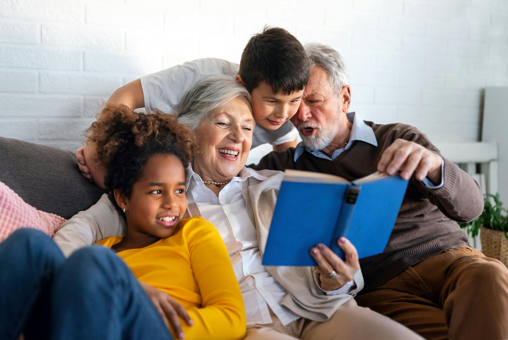 Far- och morföräldrar och barnbarn sitter tillsammans i soffan och tittar i en bok.