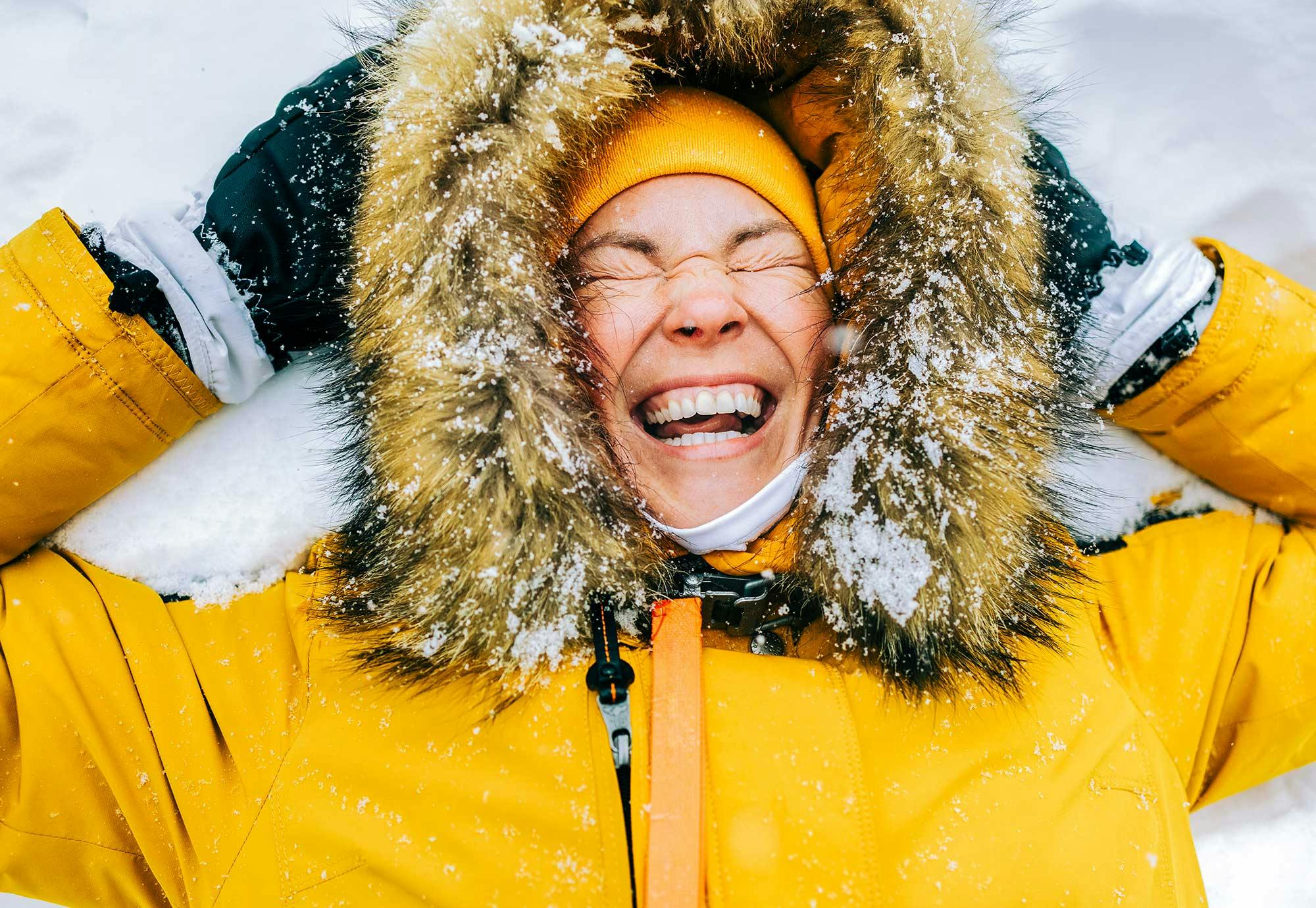 Kvinde ligger i vinterjakke i sneen og smiler.