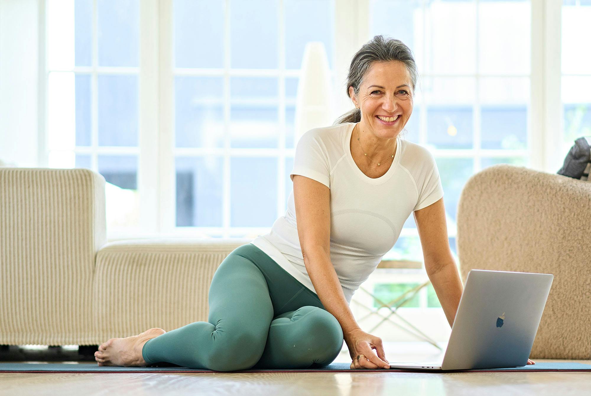 Midaldrende kvinde i træningstøj, der sidder foran en bærbar på gulvet.
