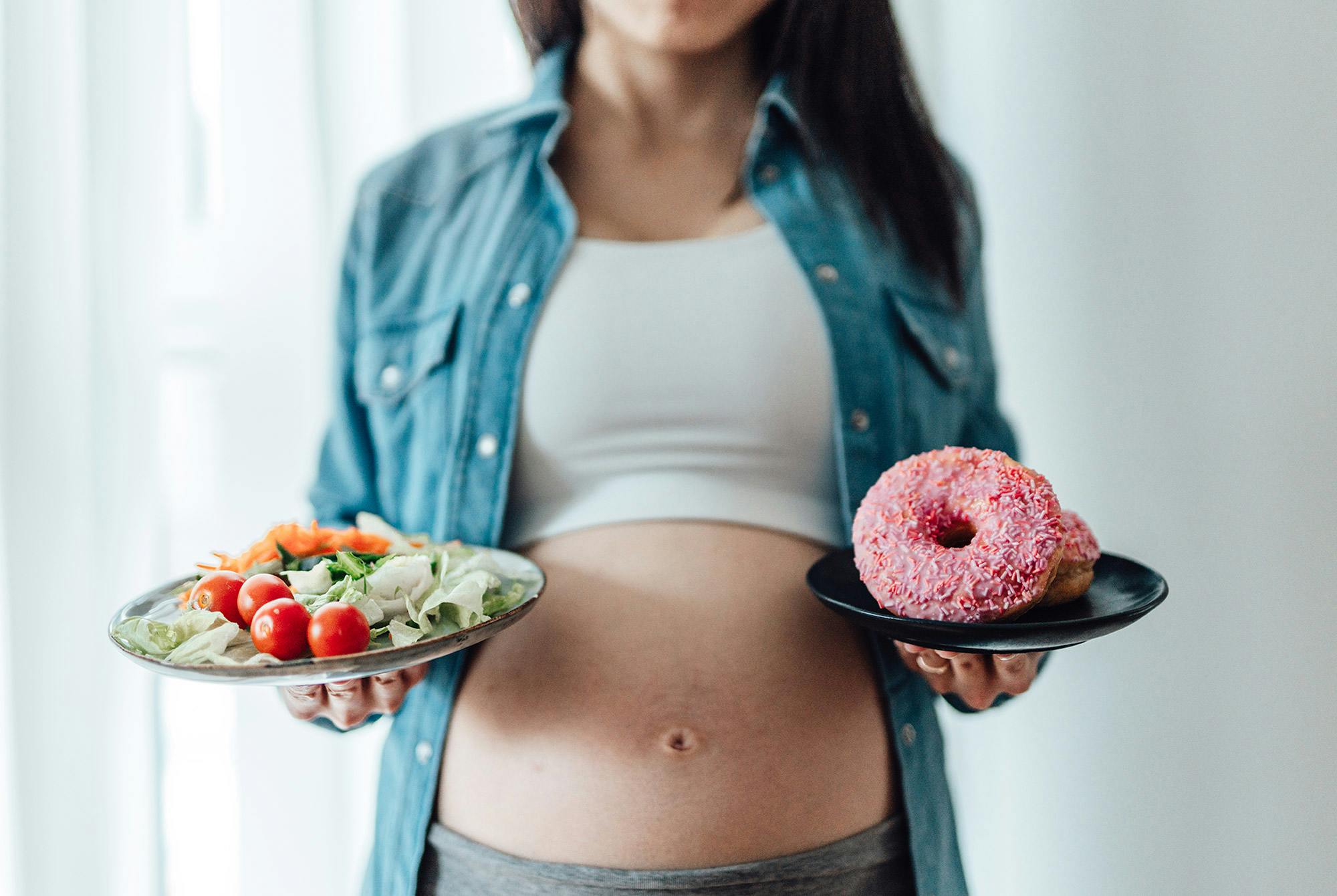 Gravid kvinne holder en salat i den ene hånden og en smultring i den andre