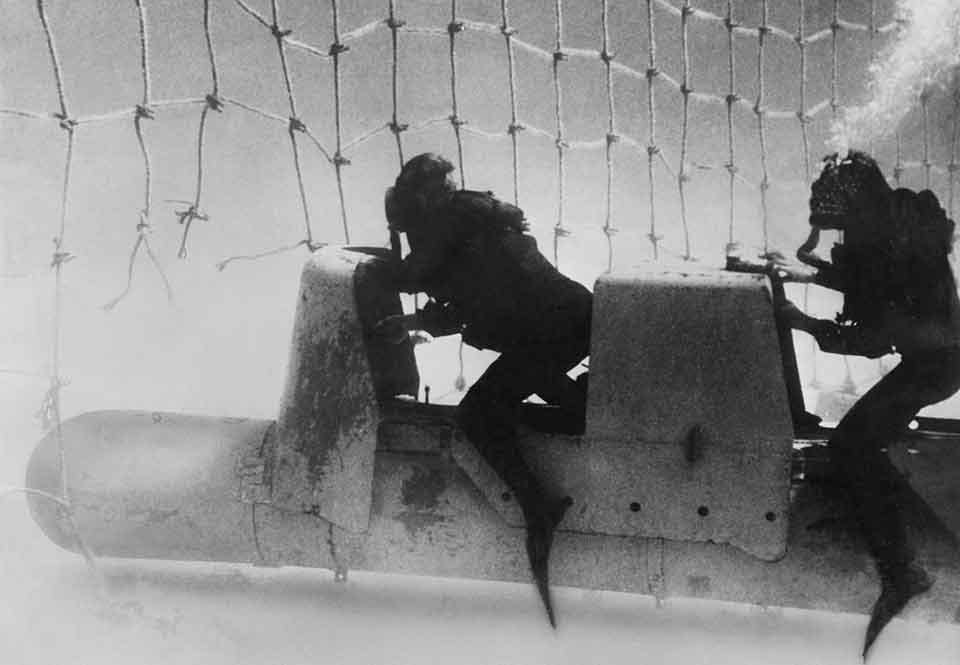 Två italienska dykare tar sig igenom ett torpednät med sin undervattensfarkost. Två italienska dykare tar sig igenom ett torpednät med sin undervattensfarkost.