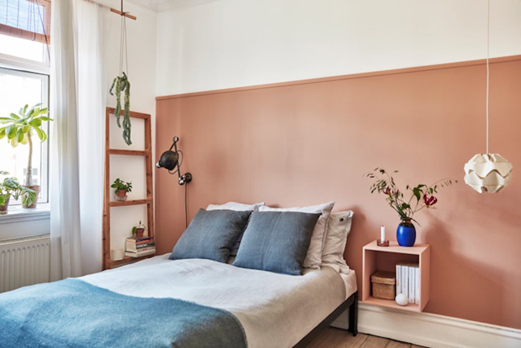 De gekleurde wand wordt ingekaderd door een simpele houten lijst. Het nachtkastje kreeg dezelfde kleur, om rust aan te brengen. De lamp links is van Jieldé, terwijl die rechts een oude Le Klint is. Het bed is van LLLP by Nicholas Rose. 