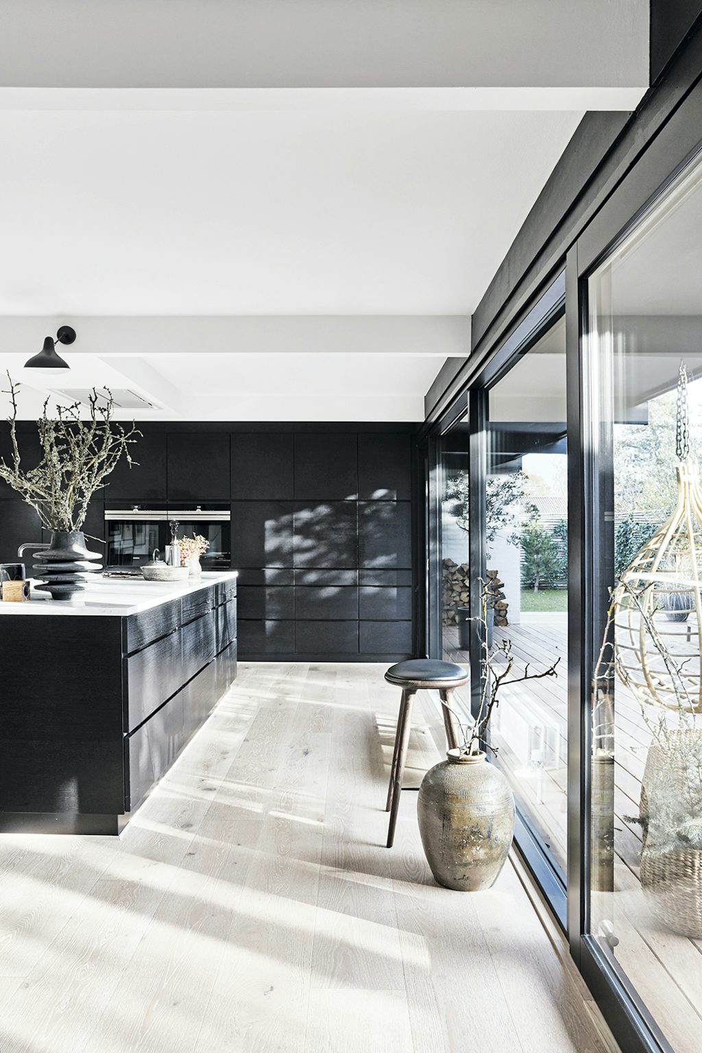 Het kookeiland van zwartgebeitst hout is van Kvik. De barkruk is een ontwerp van Space Copenhagen voor Mater en de vaas komt van Atelier du Noir. De lamp is een Mantis, ontworpen door Bernard Schottlander. Minimalistische zwarte keuken met kookeiland en barkruk.